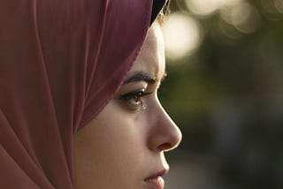 A side profile of a woman in a dusty pink hijab gazes into the distance, her expression solemn and reflective.