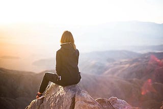 A woman sitting at the edge of a rock staring at the mountain valley with the sunset