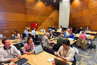 A room full of people engaged in group discussions around tables, using laptops and taking notes.
