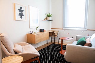 A bright therapy room with a wooden desk, pink chair, and beige couch.