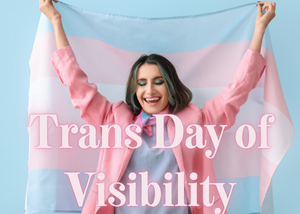 Woman holding Trans Pride Flag with great green and brown hair, a pink jacket and light blue shirt. The words Trans Day Of Visibility overlaid on top