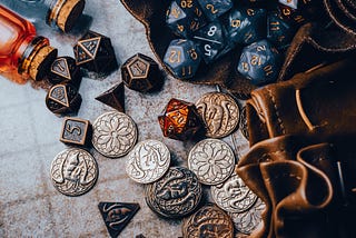 Image: A collection of scattered coins and dice spilling out from pouches and bottles. I’m looking at you, you greedy little dice-hording goblin.