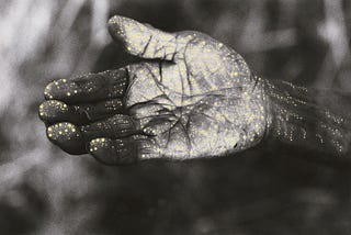 Close-up of an older indigenous woman’s hand is held out, palm uppermost. The photographed hand has been painted over with patterns of tiny white and yellow dots.