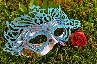 Red rose beside a blue sparkly Mardi Gras mask lying in the grass.