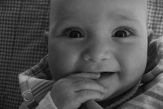 a young white skinned baby smiles directly into the camera. The baby’s eyes are sparkling with laughter. It is a close up headshot. You see the baby has their right hand in their mouth, and they are smiling but don’t yet have any teeth. They are wearing something stripy, and lying on a plaid blanket.
