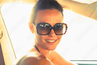 Beautiful   woman sitting in a car with big sun glasses and a black bikini on. the sun is shining on her, and her hair is pulled back into a pony tail.