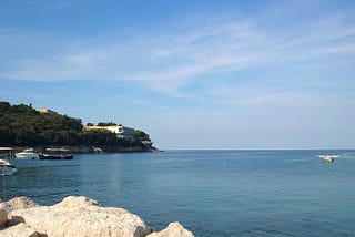 Beautiful little bay, quiet blue sea, some rocks in the foreground, a few boats in the background. A house on a green hill. Summer break. Pauza.