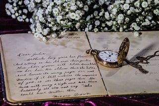 An old journal showing handwritten entry, a pocket watch and chain lies on the open pages, in the background are white flowers, baby’s breath, vintage, romantic, retro