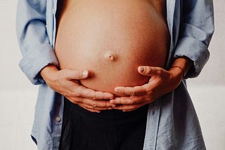 The picture shows a close up of a bare, large, pregnant stomach. The woman has both hands resting underneath and her belly button is in the center of the frame.