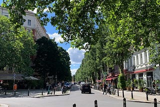 Photo of a treelined street in Berlin. It’s quiet with only few people and one car. The sky is a beautiful blue and the sun is shining.