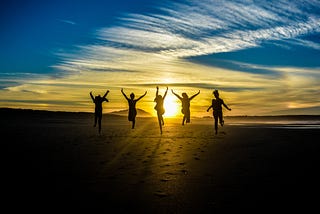 A group of people running on sand towards a sunset, leaping into the air