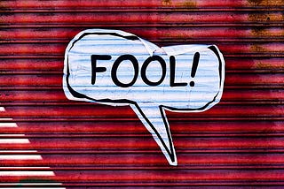 A white comic speech balloon containing the word “FOOL!” It appears to be on a red wall.