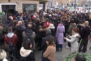 A crowd of about a hundred people, dressed in city winter clothes, crowding on the sidewalk in front of a building.