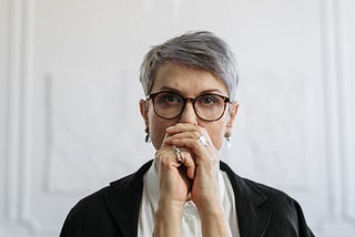 A woman in her late 50s/early 60s stairs straight at the camera looking stern with her hands folded in front of her mouth.