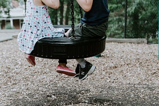 A close-up shot of two children on a tire swing. One is wearing a patterned dress and pink shoes. The other is wearing a dark navy shirt, dark green shorts, and black shoes. They are on a playground with wood chips on the ground and trees in the background
