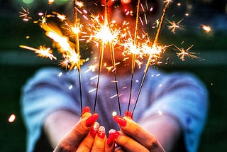 A woman with long red painted nails is holding up five lit sparklers