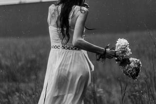 Black and white photo of a woman walking through tall grass in the rain, holding a bouquet. Her back is turned and her hair is wet, creating a moody and distant atmosphere.