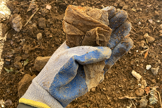 A gloved hand holds a piece of plastic waste, heavily soiled and degraded, underneath the thumb. The background is of dirt and soil in a garden.