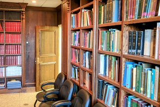Library room with tall wooden bookshelves filled with neurosurgery and neuroscience books. Left shelf holds uniform red journals. Four black office chairs line the shelves. An open wooden door leads to a hallway. Room is warmly lit and quiet.