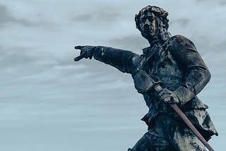 A weathered, dark-toned statue of a man pointing outward with his right arm extended.