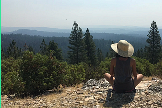 A brown-skinned woman sits on a rocky dirt area overlooking a vast expanse of evergreens. She wears a wide-brimmed hat and dark clothing.