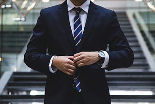 A white man in a navy suit adjusts his tie; his face is cropped