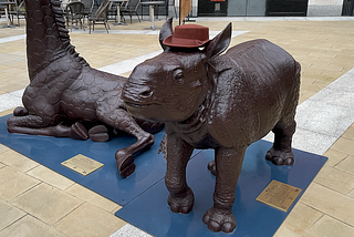 A brown statue of a Hippo calf wearing a brown porkpie hat.