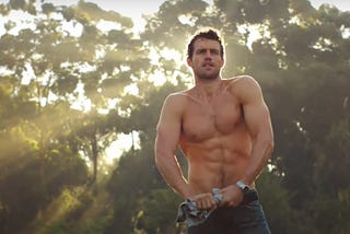 Screenshot taken from The Diet Coke Ad on You Tube. Showing a man set against trees with piercing sunshine rays, he has his top off and is ringing it.