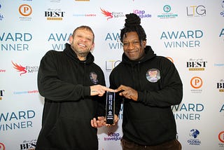 This images shows Stephen Arnott and David Okwesia from Beats Bus collecting the Hall of Fame award. They’re both holding the trophy between them infront of a background with sponsor logos