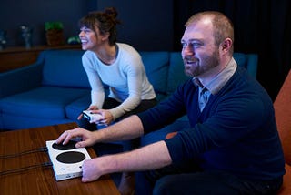 A man playing an unseen video game with a Xbox Adaptive Controller alongside a woman holding a typical Xbox controller. They are laughing and smiling at a screen out of frame.