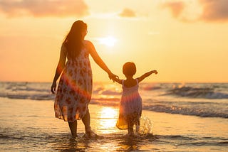 back of a mom and child walking into the sunset while wading through waves in water