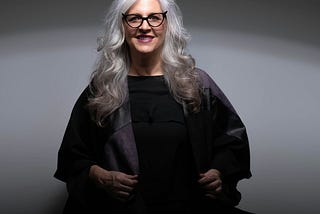 A white-haired woman with glasses is dressed in black and smiling.