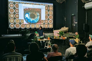 A crowd of people infront of a presentation in a dark room. The presentation shows a human iris and a pattern of circular brown images in the background. There are a couple of potted plants on the stage.