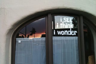 Image shows a two panes of a window with sheer white curtains covering the lower part of each; on the left, there’s a reflection of sunset and streetlights; on the right, the words. “i see i think i wonder”