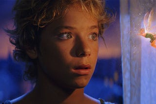 Young blonde boy, Peter Pan, looks wide eyed at the tiny glowing fairy hovering next to him, Tinkerbell, in a green dress with sparkling wings
