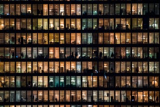 Side view of an office building seen from a distance. Brown wooden frame ceiling offices with glass windows, the silhouette of people inside. Image by Creative Lab @ scop.io