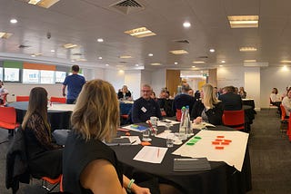 Panoramic shot of a conference room with people sat in groups at round tables. There are flipchart sheets with post-its on along with other typical conference paraphenalia