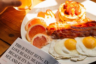 Plate of bacon, eggs, grapefruit and pancakes. Magazine opened, leaning against the plate.