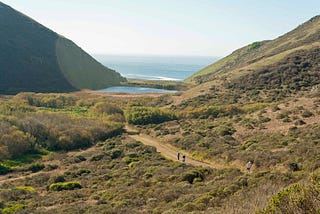 A sweeping landscape of the Marin Headlands, taken from the Tennessee Valley Trail, that includes hikers in the foreground and the Pacific Ocean in the distance.