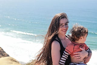 A white woman with long brown hair faces the camera smiling and holds a toddler with curly hair who faces away from the camera against the backdrop of the ocean.