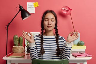 Relaxed Asian woman meditates at workplace, sits in zen pose against desktop, tries to relax after work, isolated over pink background