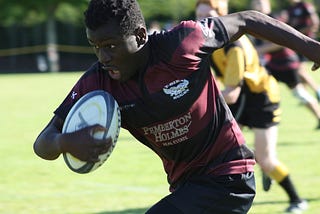 a young man runs with a rugby ball under his arm.