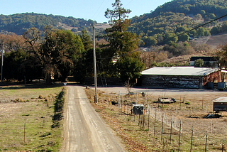 Road crossing a ranch with trees and mountains on the background a few animals to the left and a few human-made structures to the right like a hut a barn and fences. This is just a cool picture of where I live and is not really relevant for the article.