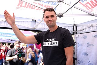 Charlie Kirk stands under an outdoor tent waving. He wears a black and white shirt with the text “ANTI-DEI PRO-MERIT”.