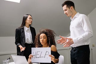 2 colleagues argue in the workplace while a lady sits in between them holding up a ‘help’ sign.
