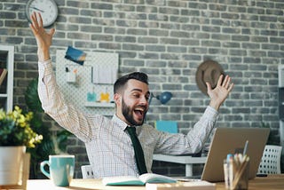A man setting at his desk has a big smile and his arms raised in the air like he’s celebrating.