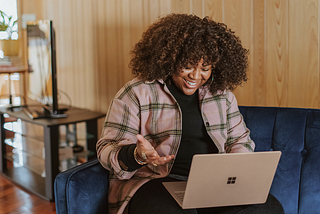 Photo by Surface on Unsplash — Woman with curly hair sitting on blue couch, smiling and gesturing to an open laptop computer on her lap