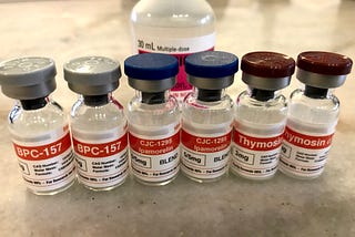 6 small, unopened peptide vials on a table with a bottle of bacteriostatic water behind them.