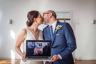 Bride and groom kiss while holding a laptop showing their Zoom wedding guests at a 2020 Covid Wedding.