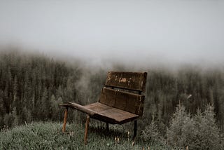 A rough-worn wooden bench sits atop a mountain hill in a forest drenched in mist.